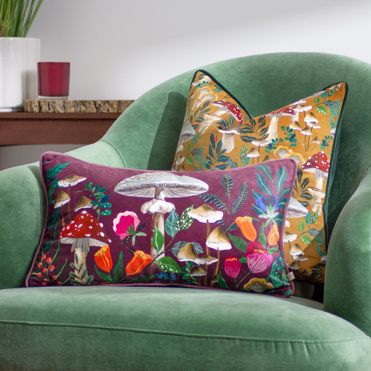 Wildgarden Mushroom polyester cushion in plum velvet with toadstool and floral embroidery, paired with an amber mushroom-print cushion on a green upholstered armchair.