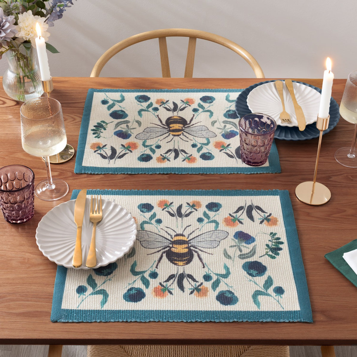 Wild Garden Animal Polyester Placemats with bumblebee and mirrored floral design in cream, teal, blue and orange, styled on a wooden dining table with plates, glassware and candles.