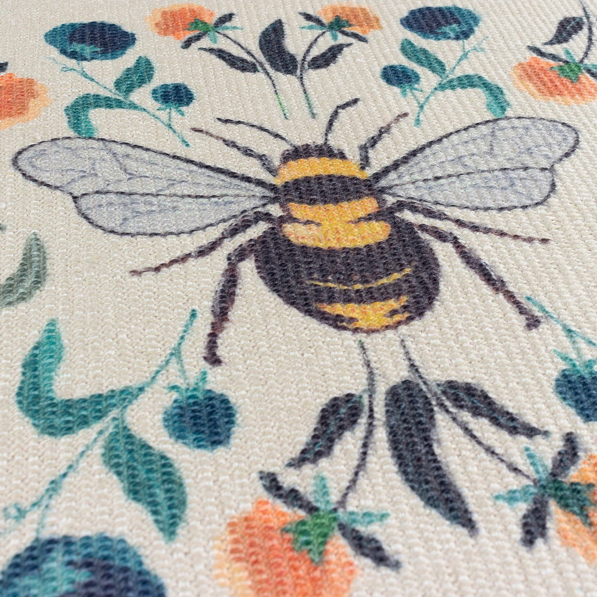 Close-up of a woven polyester placemat with a bumblebee design on a cream ground, surrounded by orange, teal and blue floral motifs and green leaves.