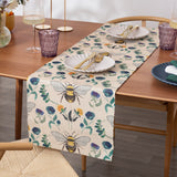 Wild Garden Table Runner in cream polyester with bumblebee and floral print, styled on a wooden dining table with white shell-shaped plates and gold cutlery.