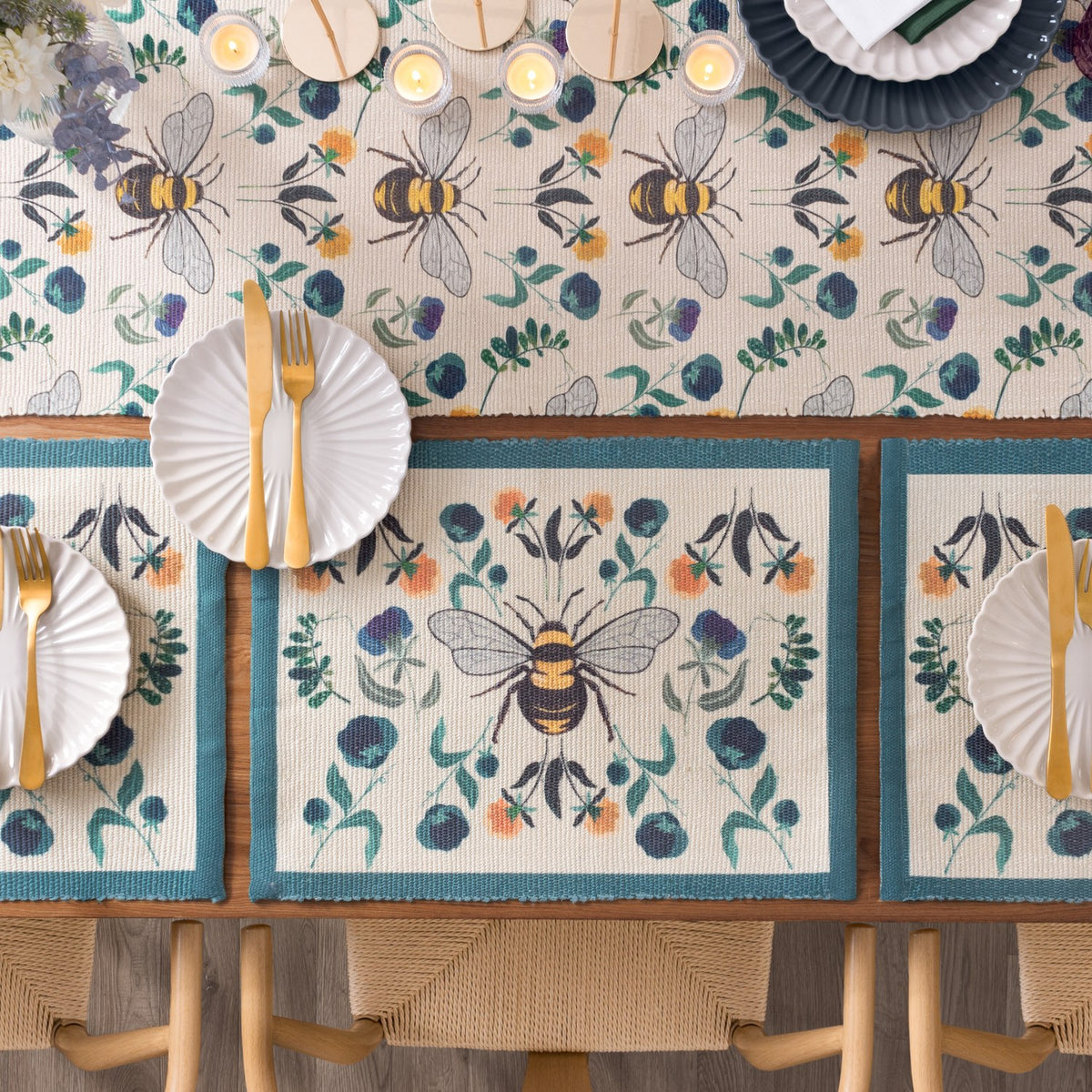 Wild Garden table runner in cream polyester with bumblebee and floral print, styled on a wood dining table with matching placemats, white plates, and gold cutlery.
