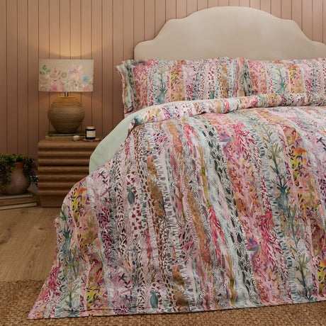 Whimsical Tale cotton duvet cover set in blush, with a vertical woodland print of trees, foliage and birds in coral, sage and grey, styled on a bed beside a lit table lamp.
