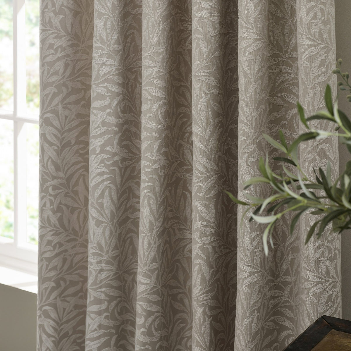 Willow Boughs Jacquard Linen pencil pleat curtains in beige, shown pencil-pleated with a woven trailing leaf pattern, styled near a window with a green plant nearby.