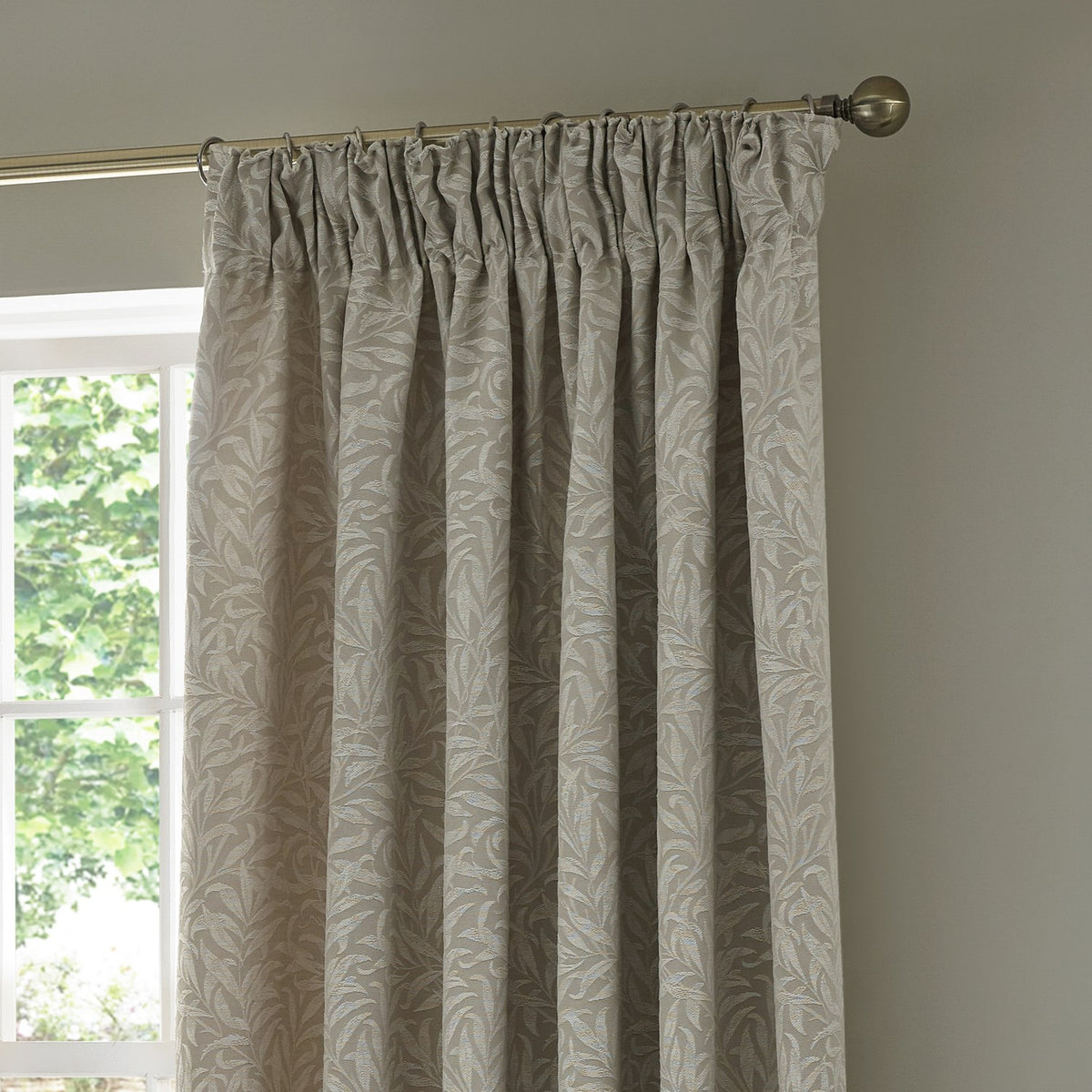 Willow Boughs jacquard linen pencil pleat curtain in beige, shown gathered on a brass pole beside a window, with woven leaf pattern in a neutral room setting.