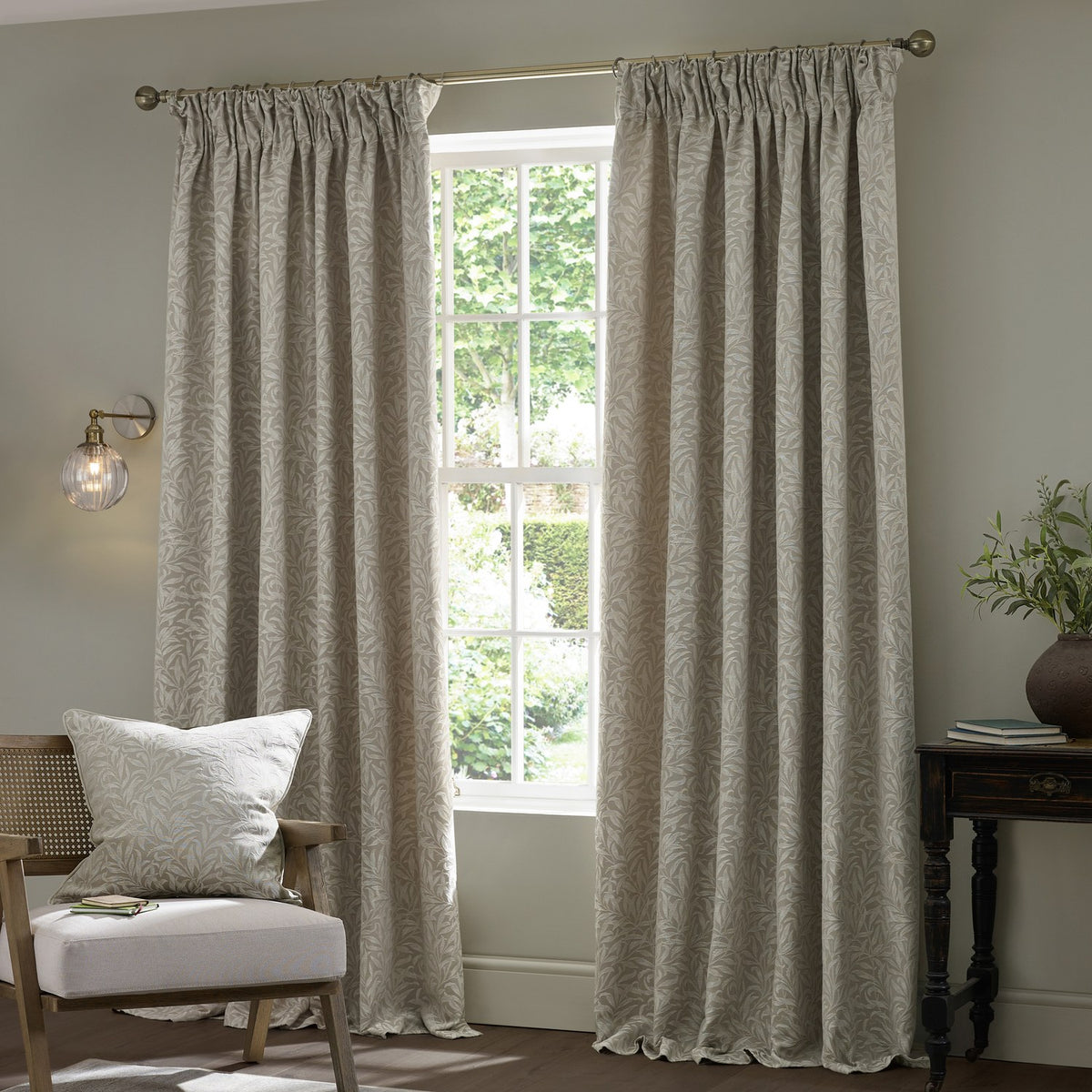Willow Boughs Jacquard Linen pencil pleat curtains in neutral beige hang either side of a window in a living room, styled with a matching printed cushion on a chair.