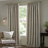 Willow Boughs Jacquard Linen pencil pleat curtains in neutral beige hang either side of a window in a living room, styled with a matching printed cushion on a chair.
