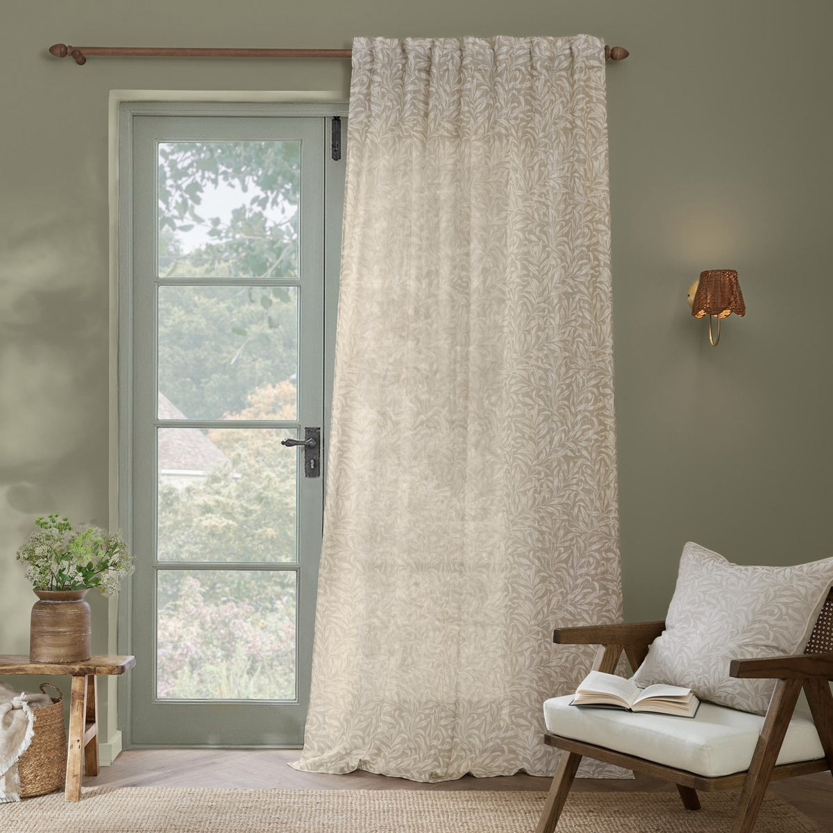Willow Bough Floral Linen curtain panel in cream with a willow leaf print, hung beside a glazed door on a wooden pole in a living room setting.