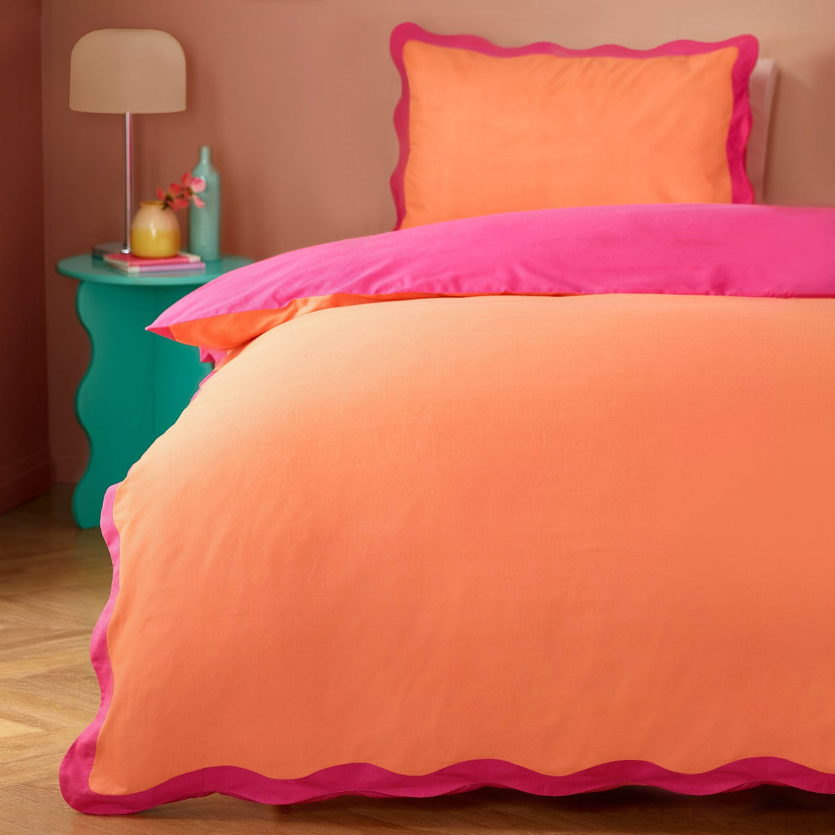 Wiggle Scallop duvet cover set on a bedroom bed, coral orange face with pink reverse and scalloped pink trim, styled with a matching pillowcase and teal side table.