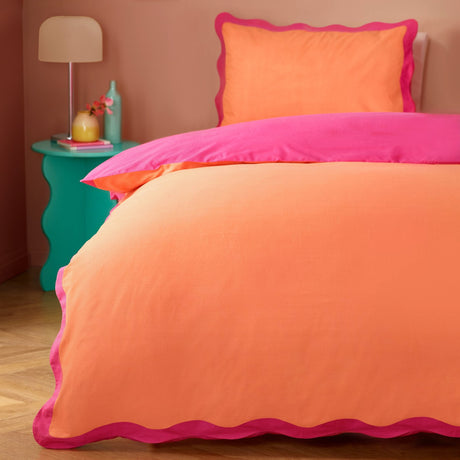 Wiggle Scallop duvet cover set on a bedroom bed, coral orange face with pink reverse and scalloped pink trim, styled with a matching pillowcase and teal side table.