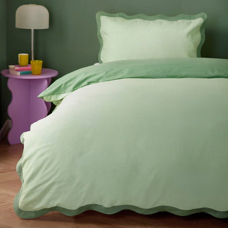 Wiggle Scallop duvet cover set on a bed in a green bedroom, pale green face with scalloped sage-green edging and matching pillowcase, styled beside a pink side table.