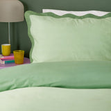 Wiggle Scallop duvet cover set in pale green with a deeper sage scalloped-edge pillowcase, styled on a bed beside a lamp and pink side table with books.