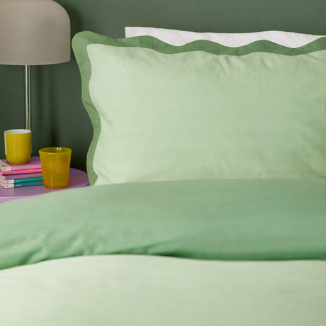 Wiggle Scallop duvet cover set in pale green with a deeper sage scalloped-edge pillowcase, styled on a bed beside a lamp and pink side table with books.