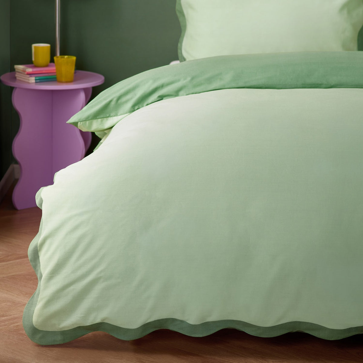 Wiggle Scallop duvet cover set in pale green with sage scalloped edges, shown on a bed beside a purple side table in a bedroom setting.