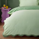 Wiggle Scallop duvet cover set in pale green with sage scalloped edges, shown on a bed beside a purple side table in a bedroom setting.
