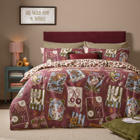 Wild Card Reversible Animal Duvet Cover Set in burgundy, showing tarot-inspired card and floral motifs on a bed, with a cream leopard-print reverse edge visible, in a bedroom setting.