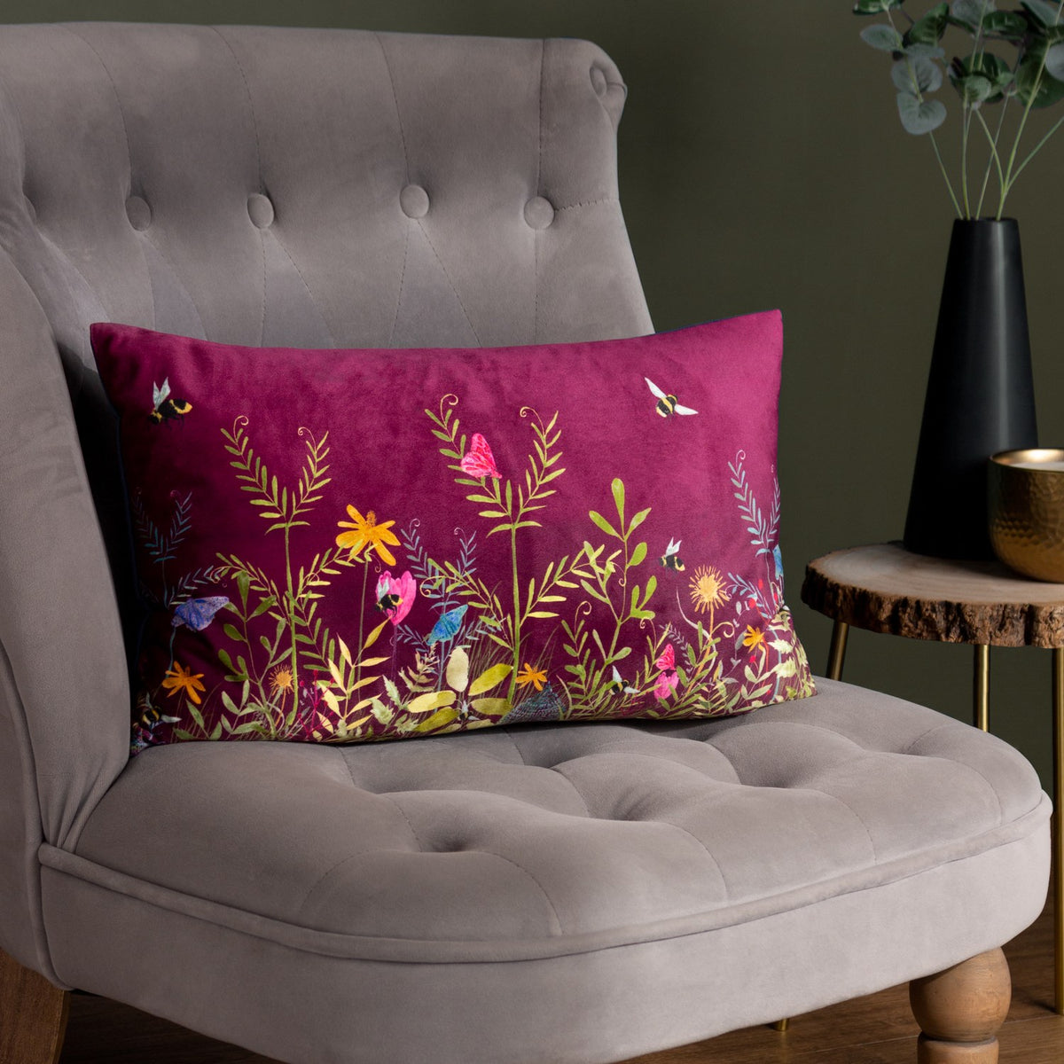 Willow Floral Velvet Cushion in Multi, rectangular bolster shape with deep fuchsia velvet front featuring floral and bee embroidery, styled on a grey buttoned armchair.