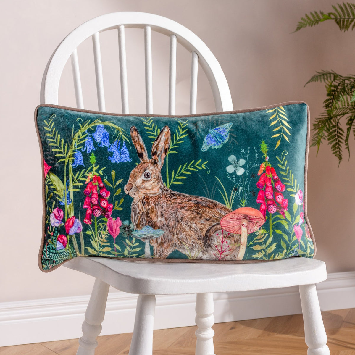 Willow Animal Velvet Cushion in multi shows a rectangular teal velvet cushion with a hand-painted rabbit among bluebells, foxgloves and toadstools, styled on a white spindle chair.