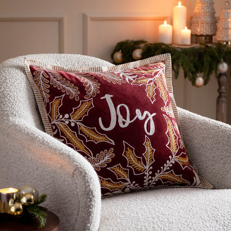 Winter Joy Velvet Cotton Cushion in red, embroidered with gold and white leaves and "Joy" script, shown on a boucle armchair beside candles and festive decor.