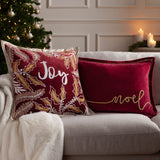 Winter Joy Velvet Cotton Cushion in red, embroidered with gold and white leaves framing "Joy" in white script, shown on a sofa beside a burgundy "noel" cushion in a festive living room.