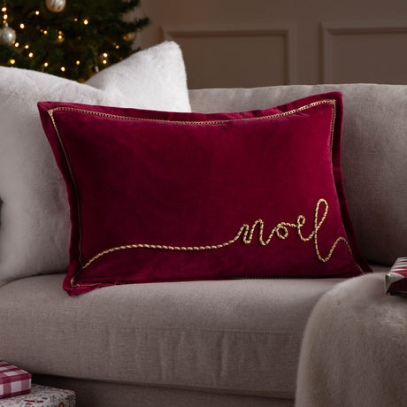 Rectangular red velvet cushion with "Noel" in cream and gold twisted cord script, gold-trimmed edge, shown on a grey sofa in a living room setting.
