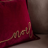 Winter Noel Velvet Cotton Cushion in deep red, close-up on 'Noel' script embroidered in cream and gold twisted cord, shown on a sofa.