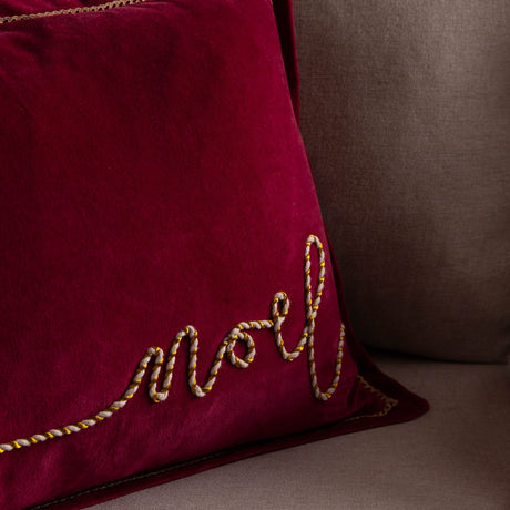 Winter Noel Velvet Cotton Cushion in deep red, close-up on 'Noel' script embroidered in cream and gold twisted cord, shown on a sofa.