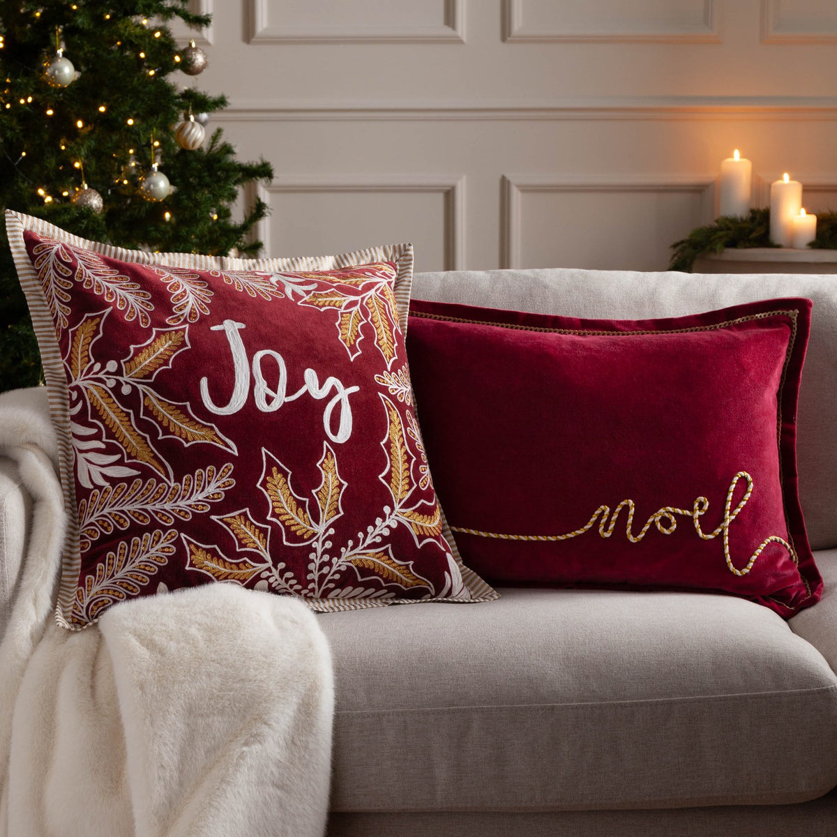 Red velvet cotton rectangular cushion with "Noel" in gold and cream rope-effect script, shown on a sofa beside a red "Joy" cushion in a festive living room setting.