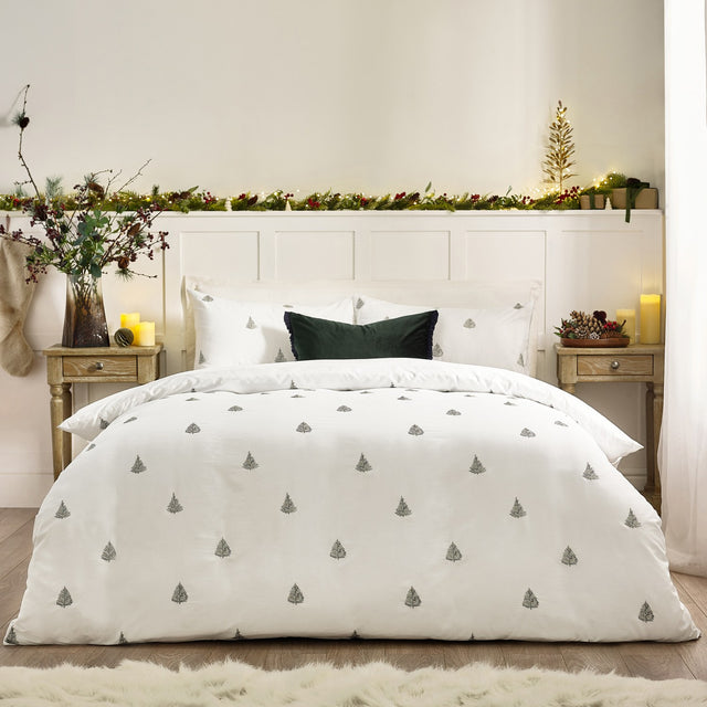 Winter Forest Woodland Duvet Cover Set in white cotton with embroidered green fir trees, dressed on a bed with matching pillowcases and a dark green cushion in a festive bedroom.