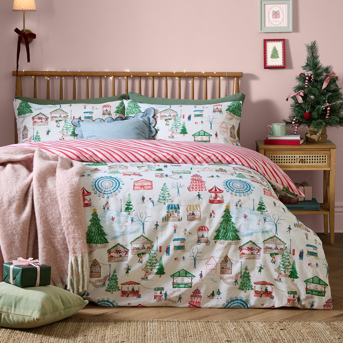 Winter Wonderland Cotton Striped Duvet Cover Set in Multi, shown on a bed with a festive fairground and market scene print, reversing to red and cream candy-cane stripes, styled in a pink bedroom.