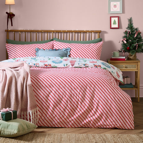 Winter Wonderland reversible cotton duvet cover set on a bed, showing red and cream diagonal candy-cane stripes with festive fairground-print pillowcases, in a pink bedroom with a Christmas tree.