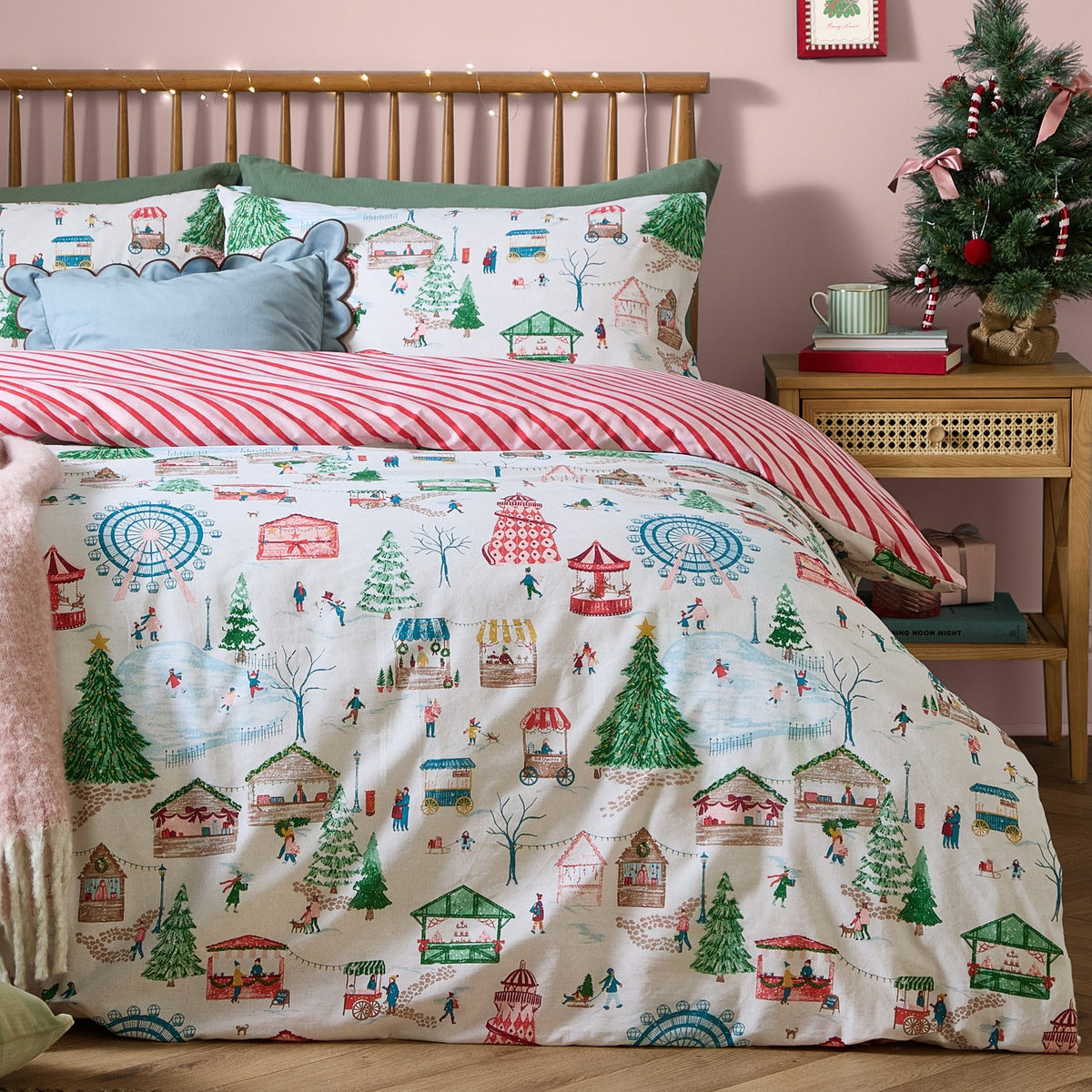 Winter Wonderland Cotton Striped Duvet Cover Set styled on a bed, showing the festive fairground print with matching pillowcases and red-and-cream striped reverse, in a pink bedroom setting.