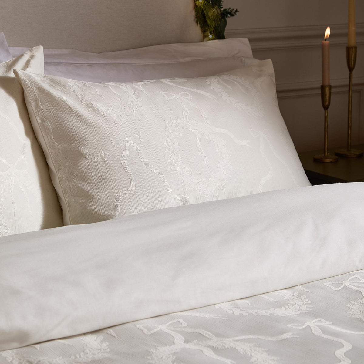 Winter Wreath Cotton Bows Duvet Cover Set in white, styled on a bed with matching pillowcases featuring a woven jacquard bow and wreath pattern, beside lit candles in a bedroom setting.