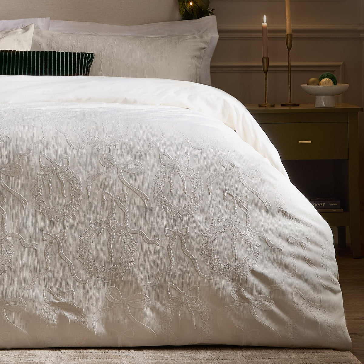 Winter Wreath Cotton Bows Duvet Cover Set in white, shown on a bedroom bed with textured jacquard bows and wreath detailing, styled with candles and a dark green cushion.