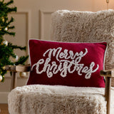 Rectangular red velvet cushion with tufted white "Merry Christmas" script and piped edge trim, displayed on a fluffy cream armchair beside a Christmas tree.