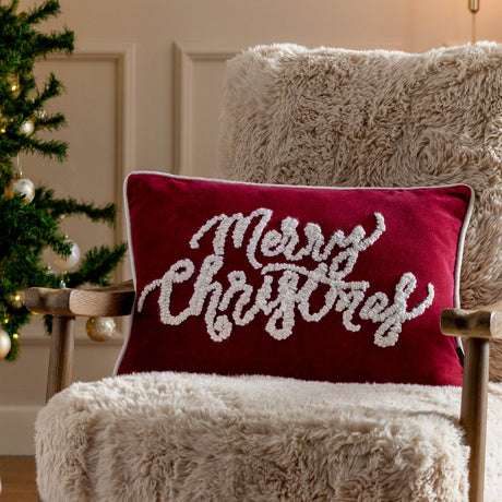Rectangular red velvet cushion with tufted white "Merry Christmas" script and piped edge trim, displayed on a fluffy cream armchair beside a Christmas tree.