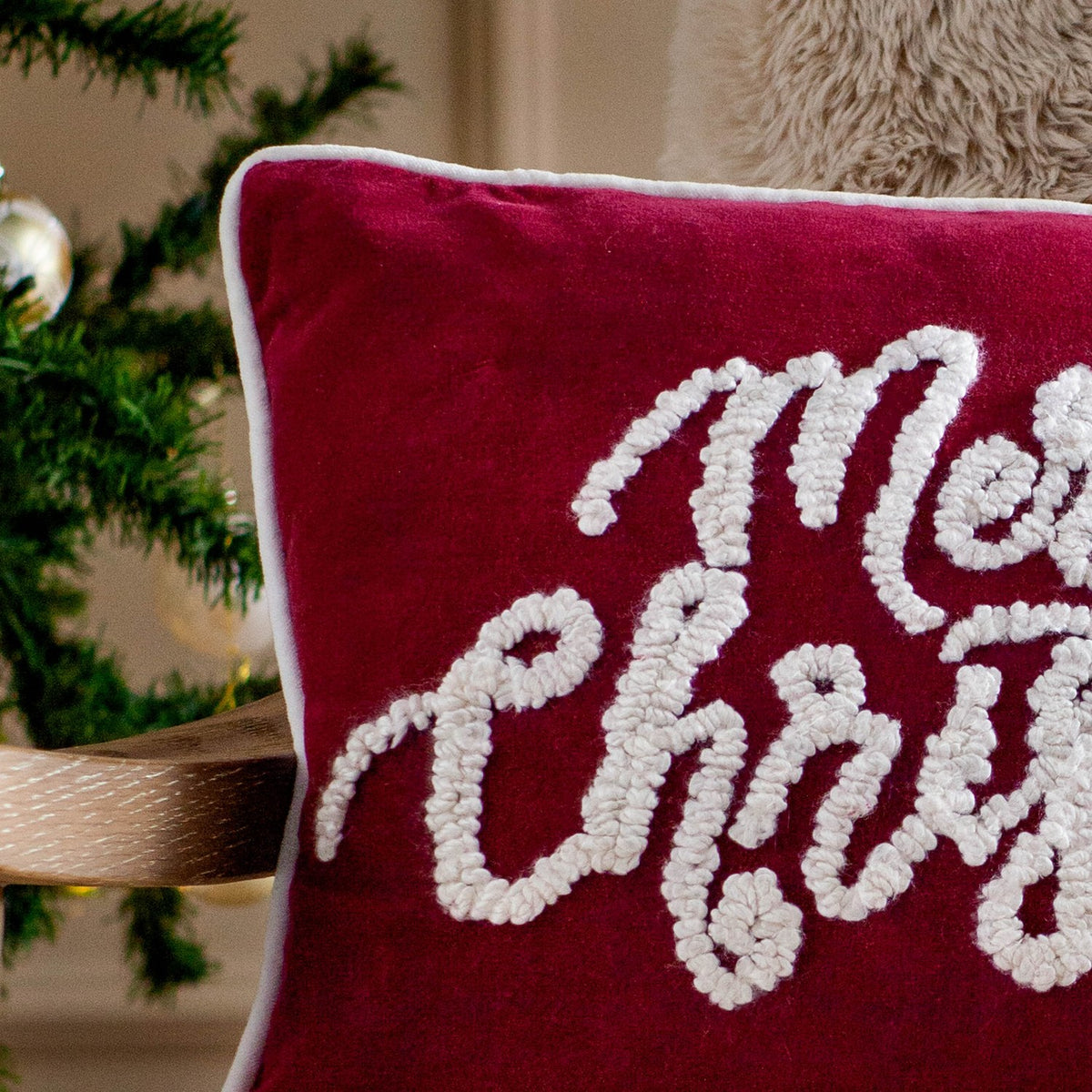 Red velvet cushion with white piped trim, featuring tufted boucle "Merry Christmas" script lettering, shown against a Christmas tree backdrop.