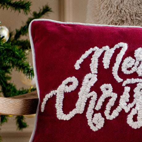 Red velvet cushion with white piped trim, featuring tufted boucle "Merry Christmas" script lettering, shown against a Christmas tree backdrop.