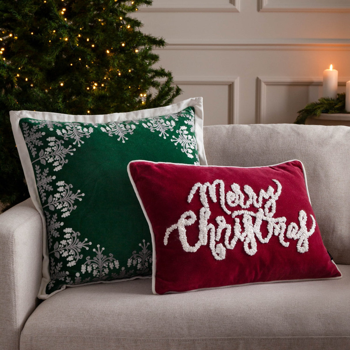 Rectangular red velvet cushion with tufted white "Merry Christmas" script and piped edging, styled on a sofa beside a green cushion and Christmas tree.