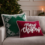 Rectangular red velvet cushion with tufted white "Merry Christmas" script and piped edging, styled on a sofa beside a green cushion and Christmas tree.