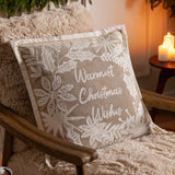 Winter Warmest Wishes velvet cotton cushion in natural beige with raised white embroidered script and floral holly design, shown on a fireside armchair.