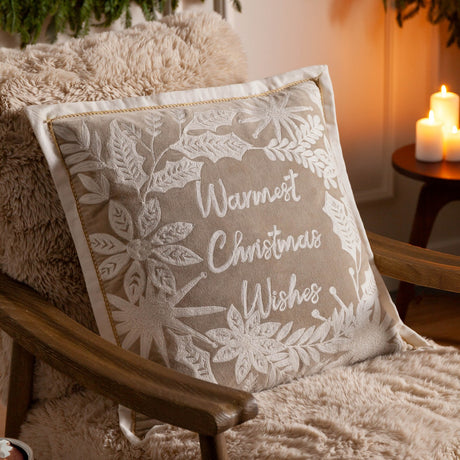 Winter Warmest Wishes velvet cotton cushion in natural beige with raised white embroidered script and floral holly design, shown on a fireside armchair.