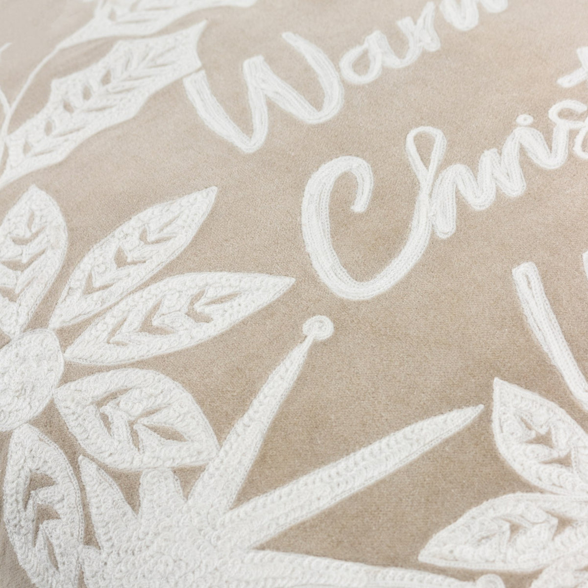 Close-up of the Winter Warmest Wishes velvet cotton cushion in natural beige, showing raised white embroidered script and leaf, holly and starburst motifs.