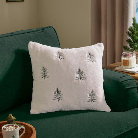 Winter Pines Woodland Faux Fur Cushion in ecru, embroidered with green and brown pine trees, shown on a green velvet sofa in a living room setting.