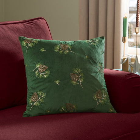 Winter Pines Velvet Woodland Polyester Cushion in spruce green with embroidered pine cone and branch detailing, shown on a red sofa in a living room setting.