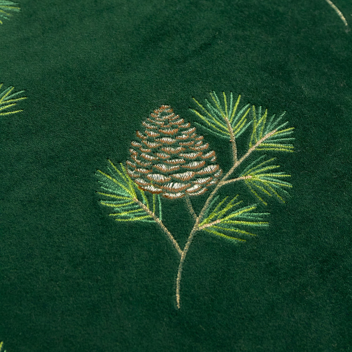 Close-up of the Winter Pines cushion in spruce green velvet, showing an embroidered pine cone and needled branch design in green and gold threads.
