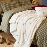 Winter Pines Woodland Polyester Throw in ecru fleece with embroidered tree motifs, draped over a green gingham bedspread beside cream pillows in a bedroom setting.