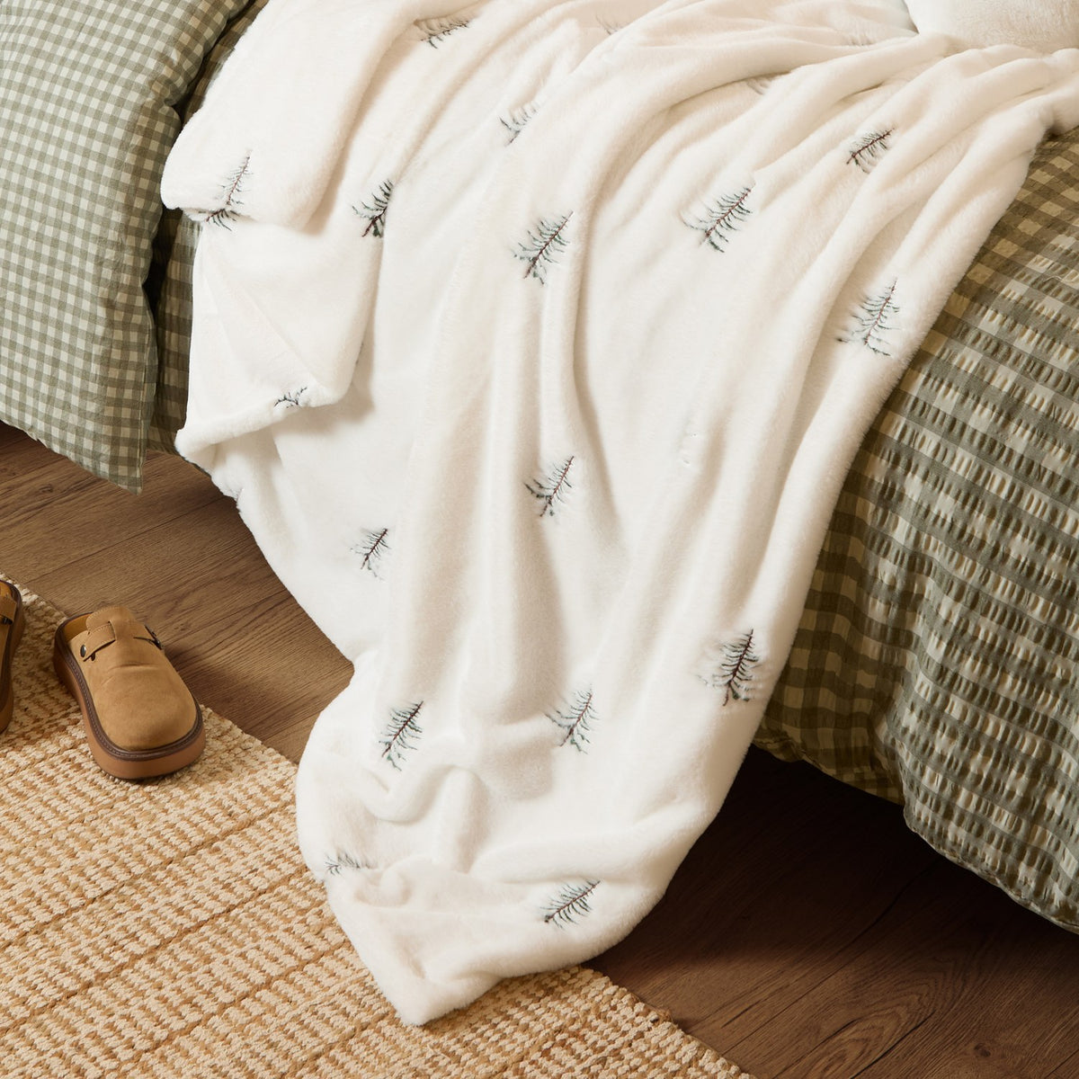 Winter Pines Woodland Polyester Throw in ecru fleece with embroidered pine trees, draped over a bed with green gingham bedding, slippers resting on a jute rug nearby.