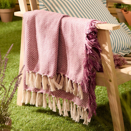 Wisley Herringbone Polyester Throw in heather/pink draped over a wooden garden chair, showing the zigzag weave with cream tassel fringe and a striped cushion behind.