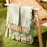 Wisley Herringbone Polyester Throw in leaf green with chevron weave and knotted tassel trim, draped over a wooden garden chair beside striped cushions on grass.