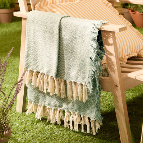Wisley Herringbone Polyester Throw in leaf green with chevron weave and knotted tassel trim, draped over a wooden garden chair beside striped cushions on grass.